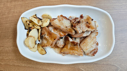 Korean Pork Belly Barbeque Grilled with Roasted Garlic on a White Plate with Light Wooden Table Background