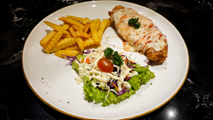 Close-up of Crispy Chicken Steak Topped with Melted Cheese Served with Crinkle Cut Fries and Fresh Salad on a White Plate with a Black Marble Table Background in a Restaurant Setting