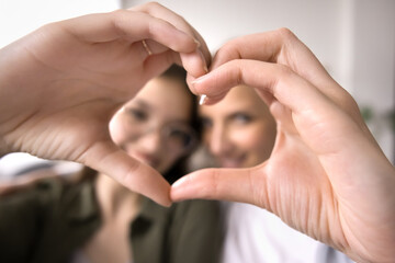 Close up face of teenager daughter and mother looking at camera, show symbol of love, demonstrate care and tenderness, enjoy time together at home, express support in family relationships. Bond, ties