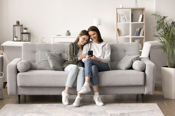 Obraz premium Attractive smiling mother relax on sofa with teenage daughter, hold mobile phone, chat in social media network, watch funny videos, learn new modern application, enjoy weekend time on internet at home