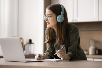 Thoughtful teen girl make assignment, wear wireless headphones studying at home, listen audio...
