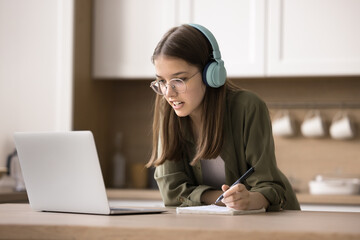 Focused teen girl in headphones watch webinar, take part in on-line study process, listens audio...