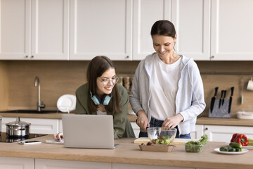 Teen studying in kitchen while her smiling mother prepare lunch for family, lead friendly talk, advising her about essay theme, help with task. Understanding, two generation communication, lifestyle