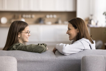 Middle-aged mom and pretty teenager daughter enjoy trustworthy conversation seated on couch, side profile view, look at each other with love and understanding, supporting, share secrets and day news