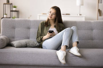 Pretty pensive teen girl in glasses sit on couch at home with smartphone, ponder on sms respond,...