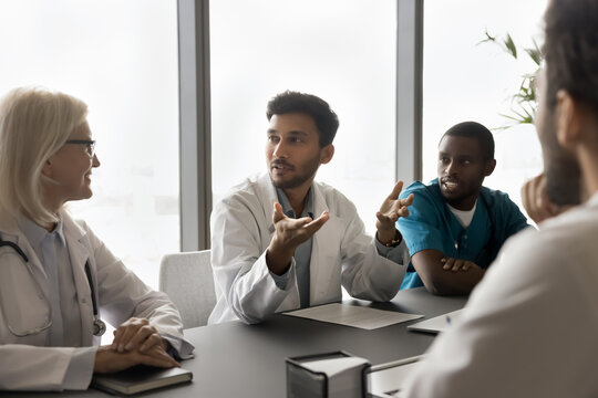 Diverse doctors listen Indian colleague, discuss treatment option, share opinion, engaged in group decision making in modern meeting room in clinic office. Seminar, medical council, knowledge exchange