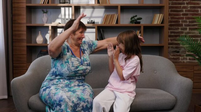 A grandmother and granddaughter are sitting on a couch at home, playing a hand-clapping game. The middle-aged woman, and the little girl are enjoying their time together.