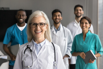 Group of diverse young and aged doctors standing in clinic office, focus on mature 60s Caucasian female general practitioner, smiling confidently, look at camera. Experience, mentorship, internship