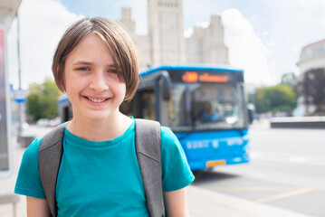 Smiling 11 year old boy with backpack waiting for bus. Teenager standing at bus stop near blue bus.