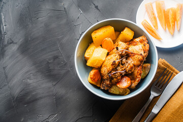 Rustic chicken with potatoes in a blue bowl on a gray background on a gray textured background with a napkin and cutlery. Copyspace.