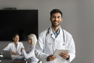 Portrait of smiling Indian male therapist, healthcare professional in white uniform holding tablet, looking at camera, standing in boardroom, two colleagues on background. Medicine, tech, profession