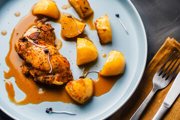 Chicken thighs baked with rustic potatoes with ju-lie sauce on a blue plate on a dark, gray background and a napkin and cutlery.