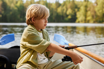 Young boy sitting in boat on lake while holding fishing rod, surrounded by peaceful nature environment with trees in background © Seventyfour