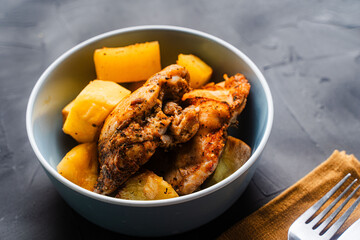 Chicken thighs baked with rustic potatoes with ju-lie sauce on a blue plate on a dark, gray background and a napkin and cutlery.
