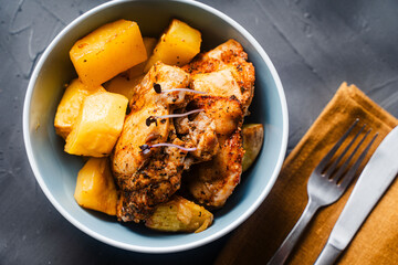 Chicken thighs baked with rustic potatoes with ju-lie sauce on a blue plate on a dark, gray background and a napkin and cutlery.
