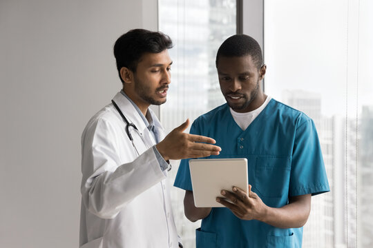 Two young multiethnic medical professionals engaged in discussion, Indian GP doctor in white coat holding tablet review patient information, data or treatment plan, explaining diagnosis to male nurse