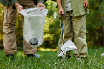 Two individuals picking up litter with trash grabber in wooded park area on sunny day, maintaining cleanliness and contributing to environmental conservation