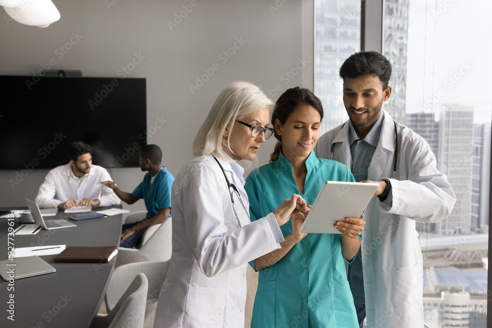 Wall mural Three diverse general practitioners in uniforms working together, holding tablet device reviewing patient data, discussing medical case, sharing knowledge, using modern technology in medical practice