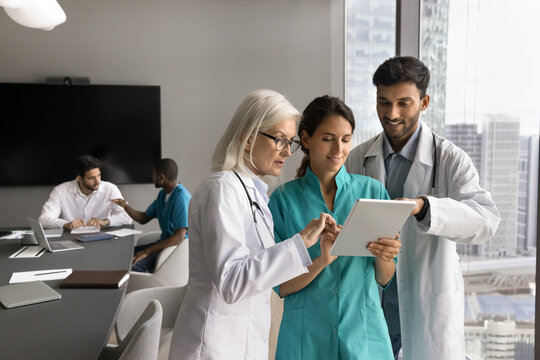 Three diverse general practitioners in uniforms working together, holding tablet device reviewing patient data, discussing medical case, sharing knowledge, using modern technology in medical practice