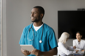 Happy thoughtful African male doctor, physician using tablet, smile, look away and thinks. Medical...
