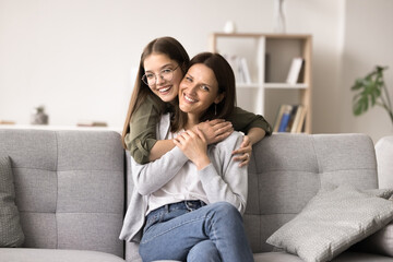 Obraz premium Cheerful middle-aged mother with adolescent daughter enjoy family weekend at home, looking at camera, pose, smile. Caring girl hugging mom from behind, express care, support and devotion, feel happy