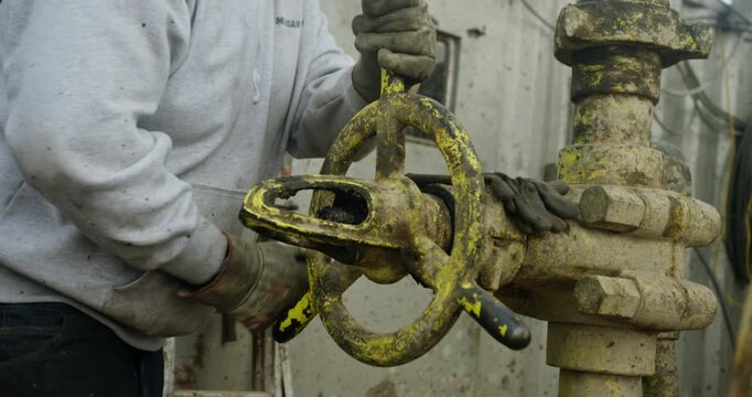 Unrecognized worker wearing dirty gloves rotates the large faucet on the oil pipe. Person at the drilling site managing the equipment.
