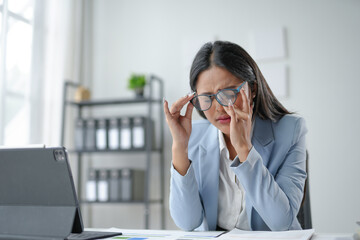 Young businesswoman experiencing a headache, removing her glasses and rubbing her eyes while working in the office