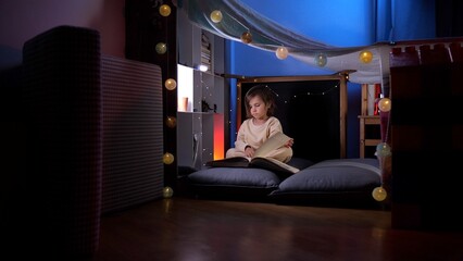 Little girl reading a book in a blanket fort at night