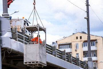 worker is suspended in a metal basket, hoisted by a crane to repair a bridge. This scene embodies the blend of human skill, engineering, and the risk inherent in construction work