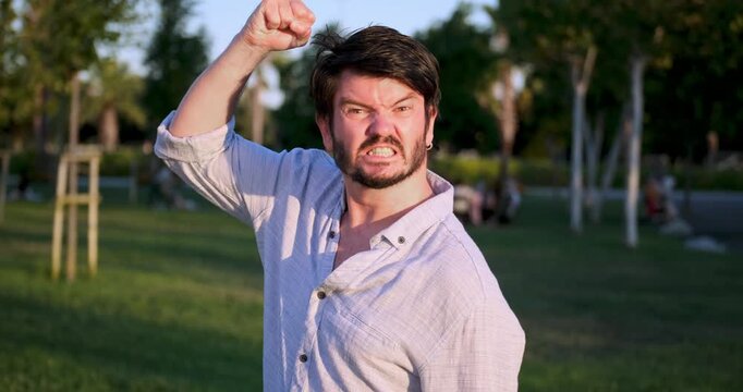 A middle-aged, dark-haired businessman wearing a shirt is looking at the camera with an angry expression in a sunny, tree-filled park on a summer day