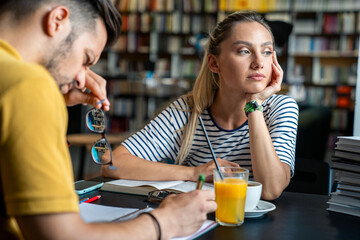 A male and female student in casual attire engage with textbooks at a library table, sipping coffee and orange juice during their study session.