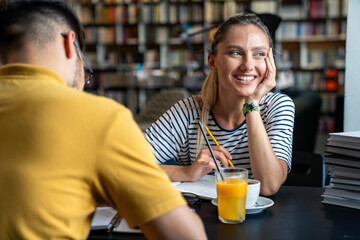 A male and female student sit at a library table surrounded by books, engaging in discussion over drinks, with the female smiling and focused.