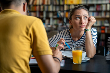 A male and female student exchange ideas in a library setting with books open, while sipping coffee and orange juice, engaged in academic discussion.