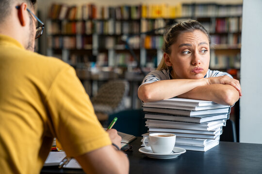 A pensive Caucasian woman rests her chin on a high pile of books, sipping coffee, as she studies with a male companion in a library setting, both clad in casual attire. - Powered by Adobe