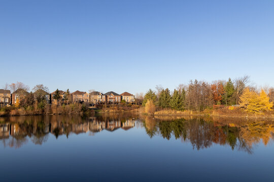 Autumnal suburban landscape with reflections in a calm lake, flanked by trees and modern houses under a clear blue sky. Taken in Toronto, Canada.
