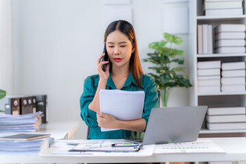 Overwhelmed and Overworked: Stressed Asian businesswoman juggles a demanding phone call and paperwork amidst a cluttered office desk. 