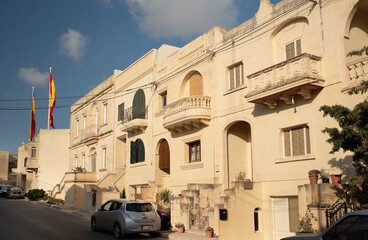 Typical picture of several houses in Malta with the typical balconies. The houses are typically light brown in colour. The picture shows a street in the city.