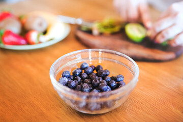 Bowl with fresh blueberries for smoothie. Overweight woman in the kitchen cutting fruit on cutting board.