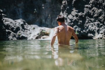 Man enjoying a swim in a natural pond with rugged cliffs in the background on a sunny day.