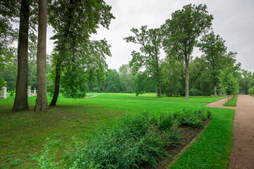 A serene walkway winds through a sprawling park, bordered by vibrant greenery and majestic trees under a cloudy sky.
