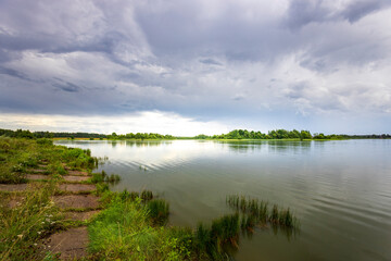 A tranquil river stretches out before a shore lined with lush green grasses and a few trees, with a cloudy sky overhead.