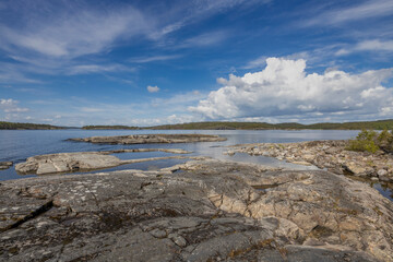 A rocky shoreline with several tidal pools reflecting the clear blue sky and fluffy white clouds. The water is calm and inviting, showcasing the serenity of the Finnish Archipelago.