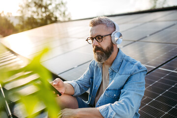 Man sitting on roof full of solar panels, headphones on head, holding smartphone.