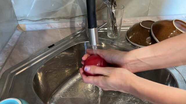 Hands thoroughly washing a red apple under running tap water in a kitchen sink with bowls in the background