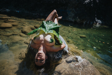 Person laying on a rock with a large leaf, relaxing in a peaceful natural environment by a clear river.