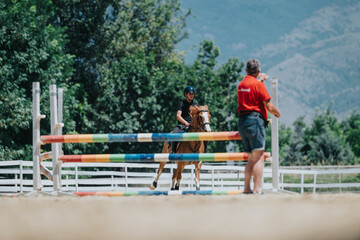 Fototapeta na wymiar Trainer guiding young equestrian as they perform a jump over colorful obstacles in an outdoor horse riding lesson in a picturesque setting.