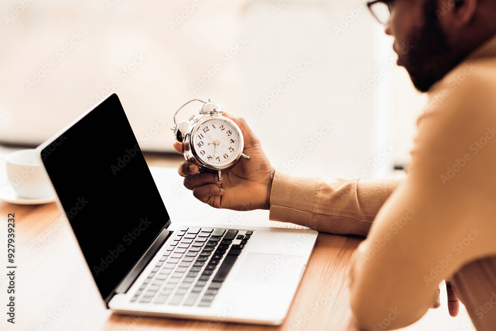 Poster deadline concept. black student holding alarm clock, working on laptop. empty space, mock up