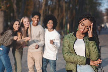 Upset Teenage Afro Girl With Multiracial Friends Gossiping And Pointing In Background, bullying...