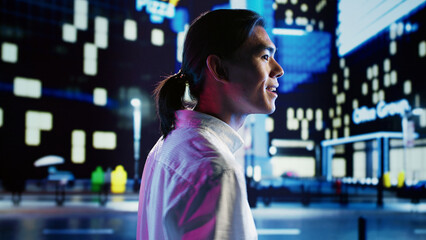 Asian man wandering around city streets at night, looking upwards to surrounding buildings in metropolitan urban environment. Smiling businessman leaving work, taking a walk outdoors