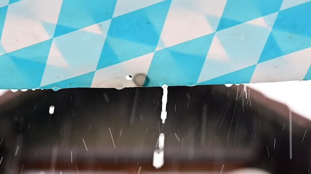 Slow motion, rain dripping from an awning that has a pattern with the Bavarian blue and white diamonds